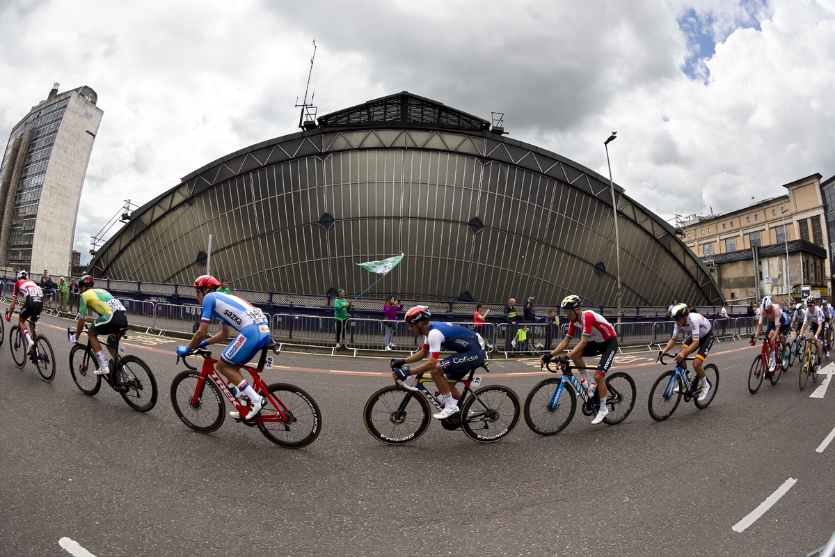 2023 UCI Cycling World Championships - Road Race - Elite Men - A group of riders pass the glass arch of Glasgow Queen Street Station while a fan waves a flag in the background