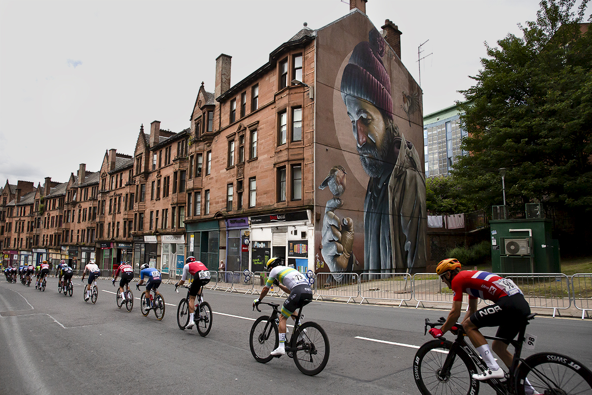 2023 UCI Cycling World Championships - Road Race - Elite Men - A strung out peloton passes a large mural of Saint Mungo painted on the side of a building