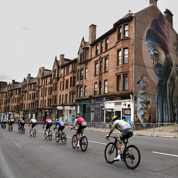 2023 UCI Cycling World Championships - Road Race - Elite Men - A strung out peloton passes a large mural of Saint Mungo painted on the side of a building