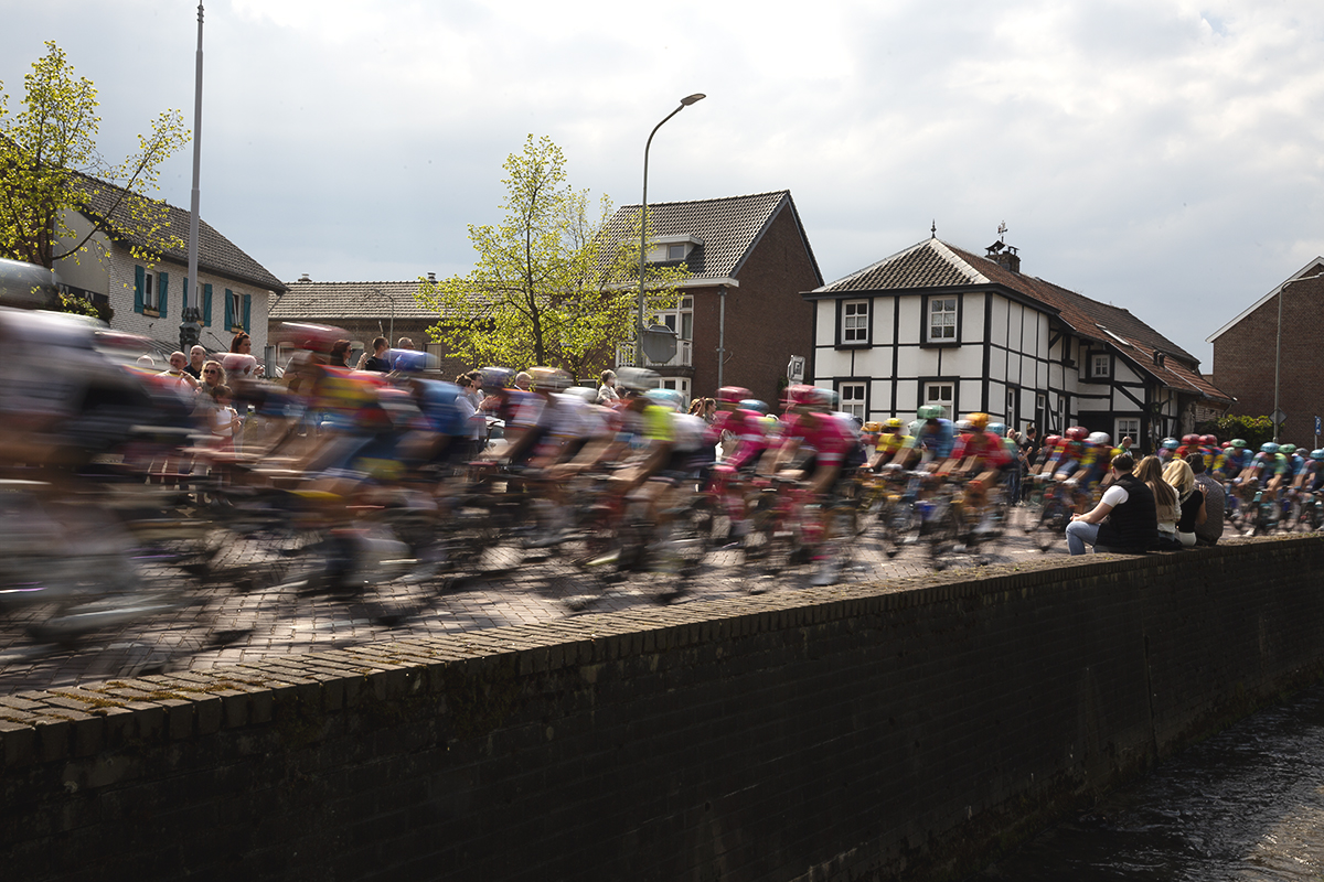 Amstel Gold Race 2025 - Spectators sit on a wall close to the road as riders speed by in Mechelen