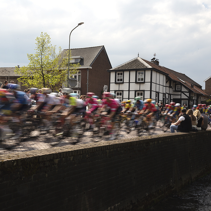 Amstel Gold Race 2025 - Spectators sit on a wall close to the road as riders speed by in Mechelen