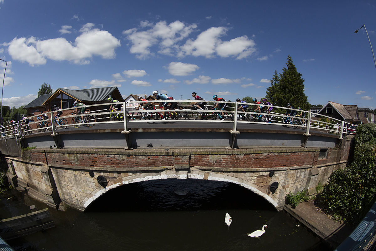 British National Road Championships 2019 -The peloton seen from the side as they cross abridge over the Norfolk Broads while swans swim by