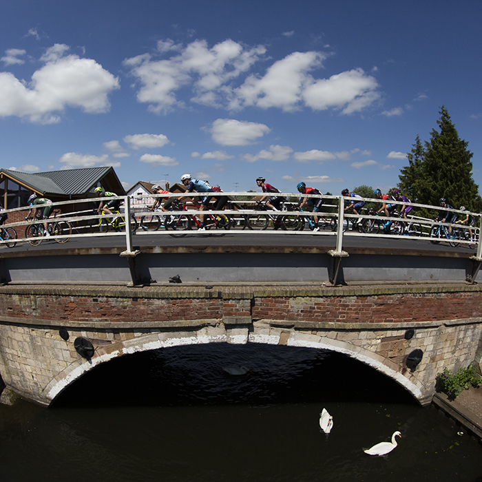 British National Road Championships 2019 -The peloton seen from the side as they cross abridge over the Norfolk Broads while swans swim by