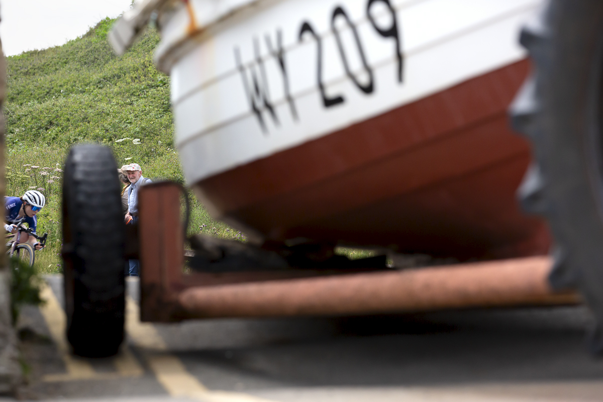 British National Road Championships 2023 - Women’s Road Race - One of the riders descends the hill framed by the fishing boat watched by an old couple