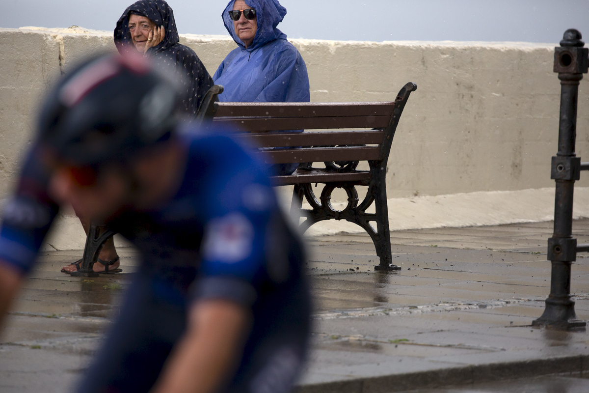 British National Road Championships 2023 - Men’s Road Race - Two people wearing rain coats watch a rider pass from a bench on the sea front