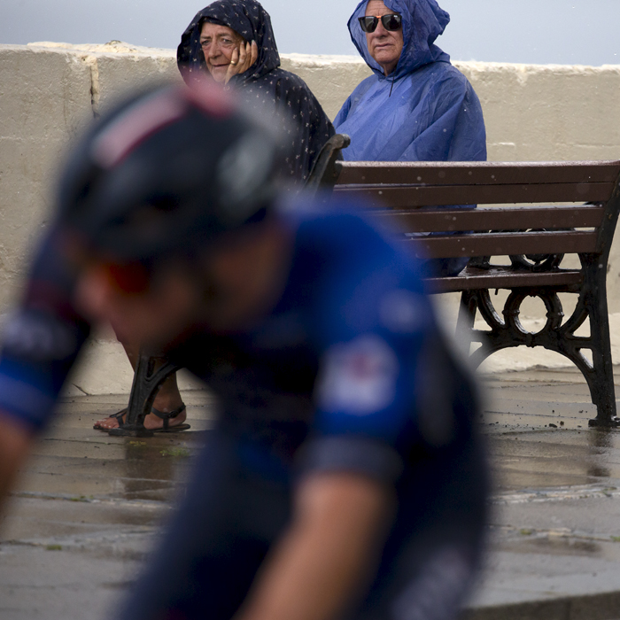 British National Road Championships 2023 - Men’s Road Race - Two people wearing rain coats watch a rider pass from a bench on the sea front