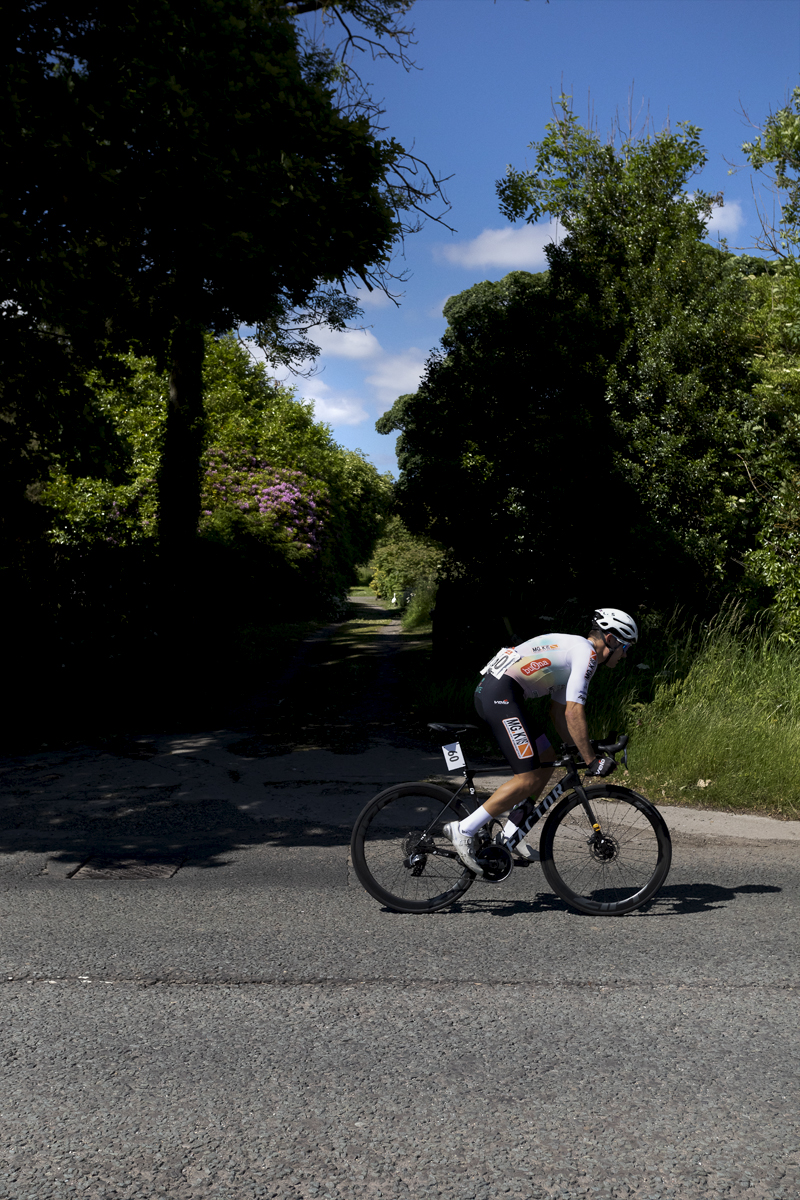 British National Road Championships 2024 - Men’s Road Race - Matthew Kingston passes a woodland track with pink flowers blooming in the hedgerow