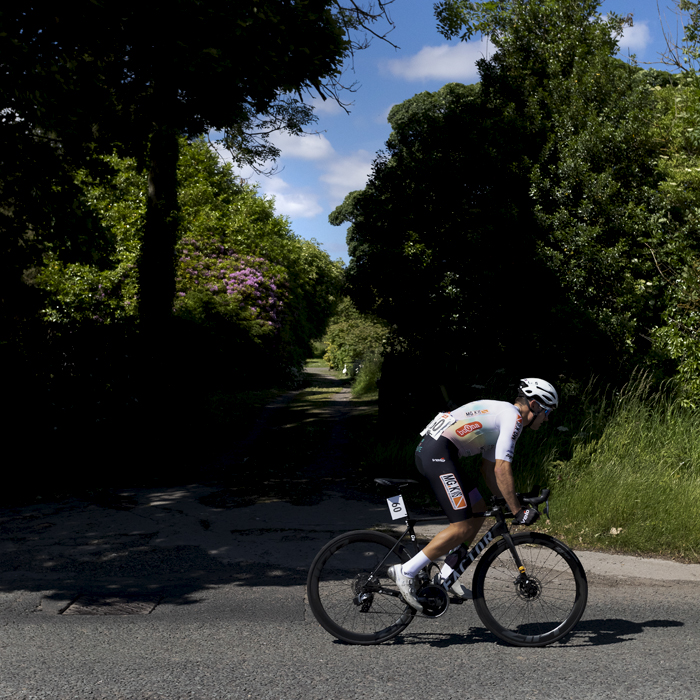 British National Road Championships 2024 - Men’s Road Race - Matthew Kingston passes a woodland track with pink flowers blooming in the hedgerow