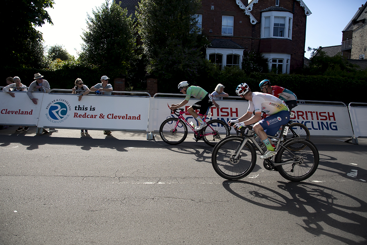 British National Road Championships 2024 - Men’s Road Race - Riders pass advertising hoardings on their way to the finish line as fans watch on