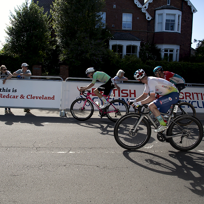 British National Road Championships 2024 - Men’s Road Race - Riders pass advertising hoardings on their way to the finish line as fans watch on