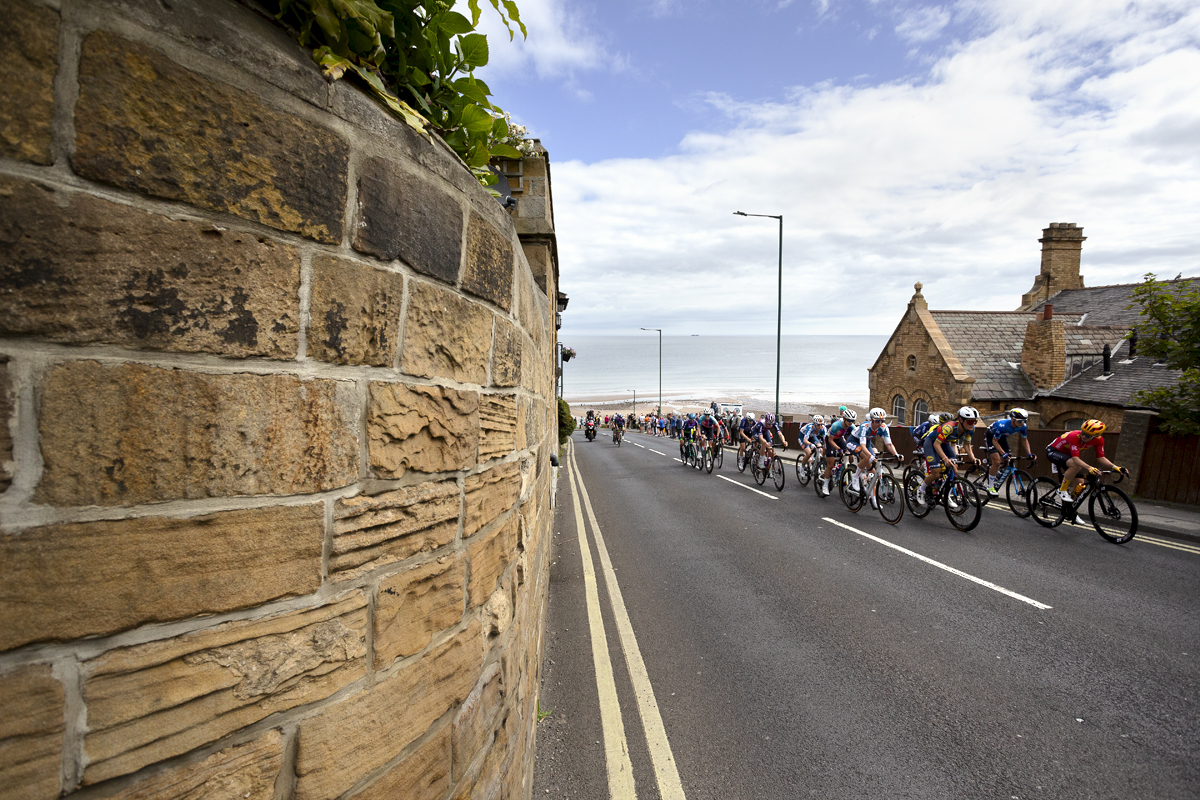 British National Road Championships 2024 - Women’s Road Race - The peloton climbs the hill past a golden stone wall with the sea and beach in the background