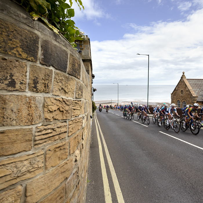 British National Road Championships 2024 - Women’s Road Race - The peloton climbs the hill past a golden stone wall with the sea and beach in the background