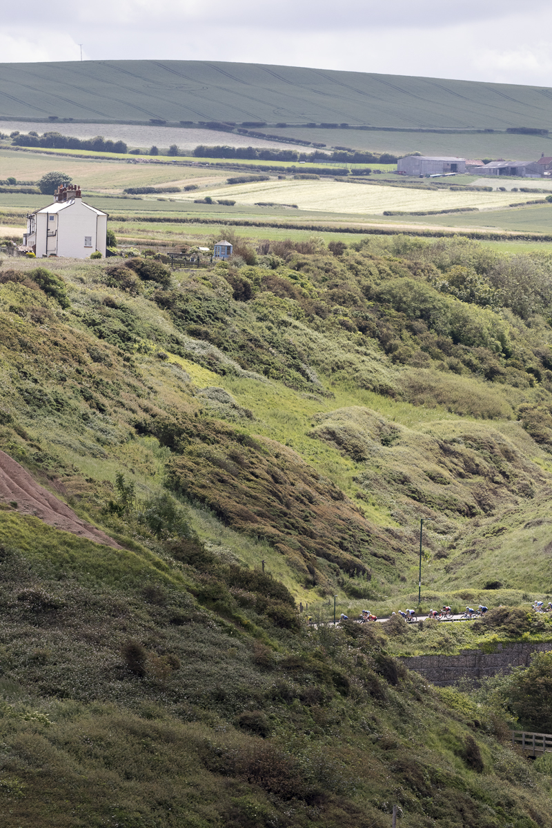 British National Road Championships 2024 - Women’s Road Race - Riders pass along the bottom of a steep sided valley with a white house on its top on the way into Saltburn