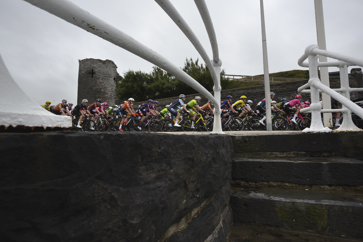 British National Road Championships 2025 - Road Race - Men - Aberystwyth - the peloton passes the castle seen from steps leading down to the sea