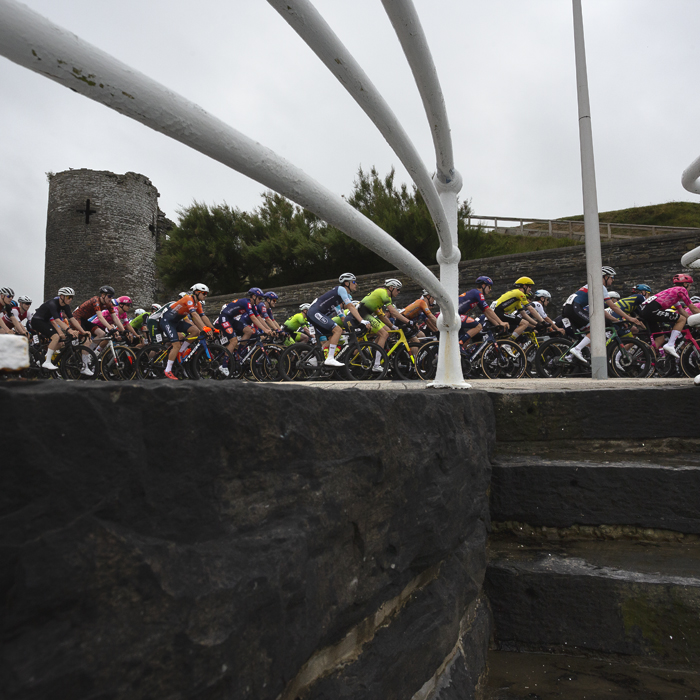 British National Road Championships 2025 - Road Race - Men - Aberystwyth - the peloton passes the castle seen from steps leading down to the sea