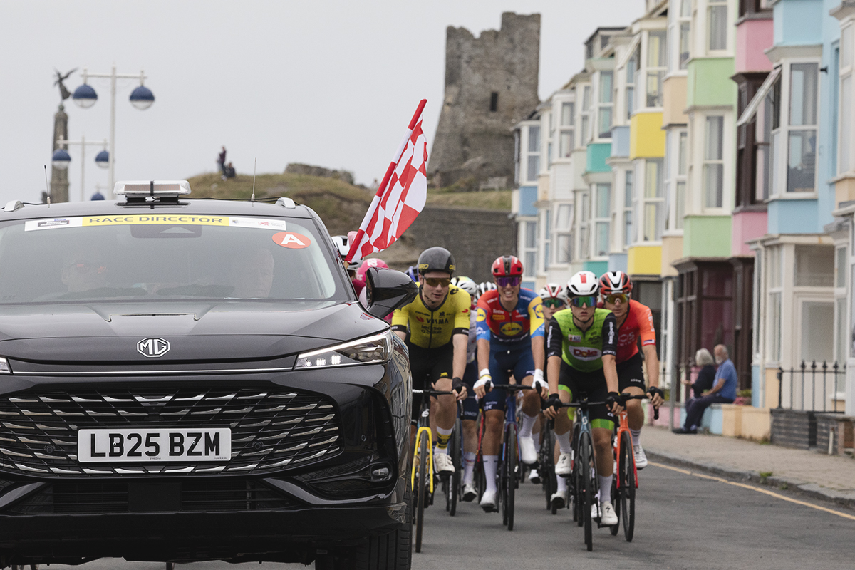 British National Road Championships 2025 - Road Race - Men - Aberystwyth - the Race Director’s car with a chequered flag hanging out the window as the riders roll out past brightly coloured houses, castle in the background