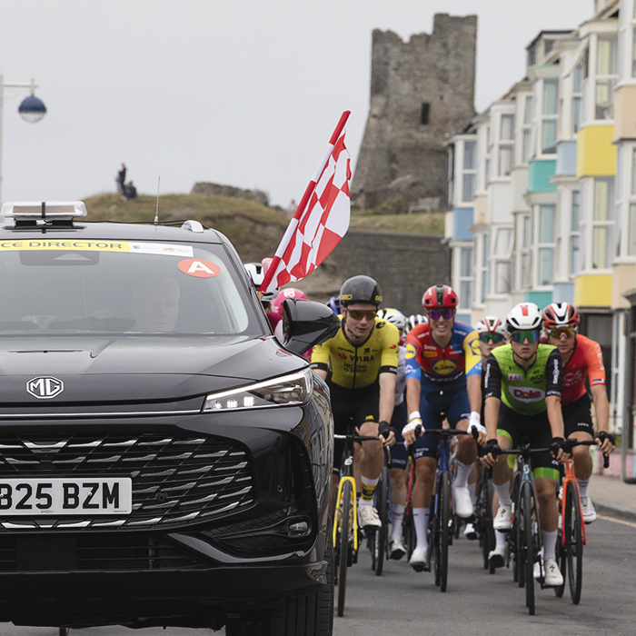 British National Road Championships 2025 - Road Race - Men - Aberystwyth - the Race Director’s car with a chequered flag hanging out the window as the riders roll out past brightly coloured houses, castle in the background