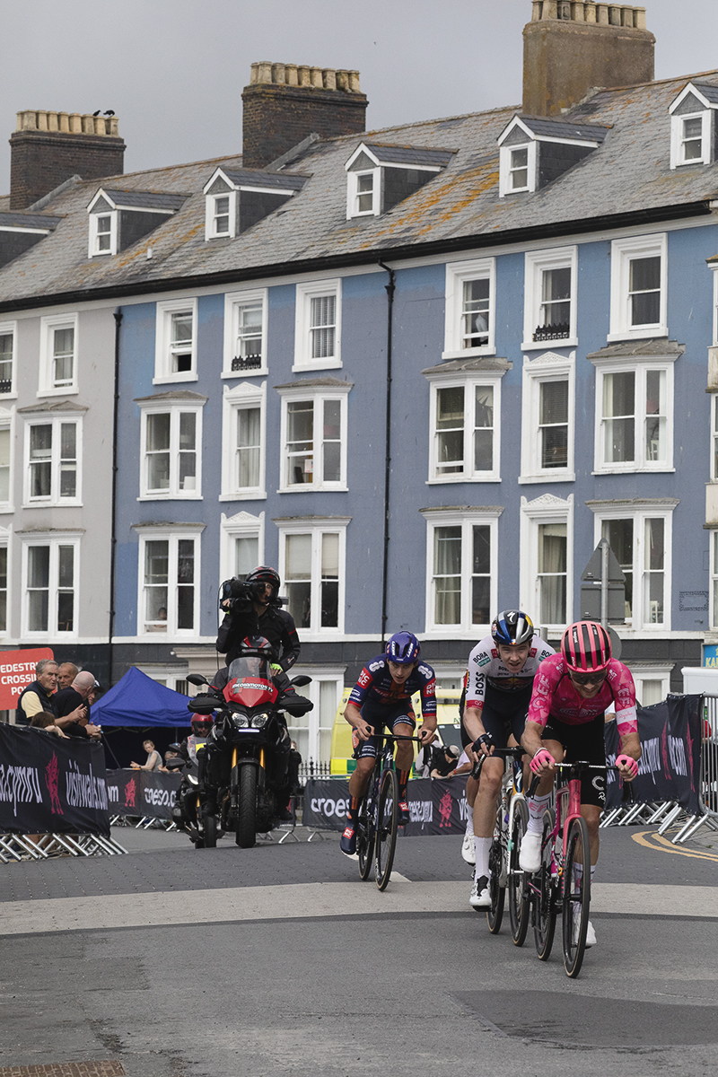 British National Road Championships 2025 - Road Race - Men - a group of riders pass houses painted in differing shades of blue on the seafront in Aberystwyth