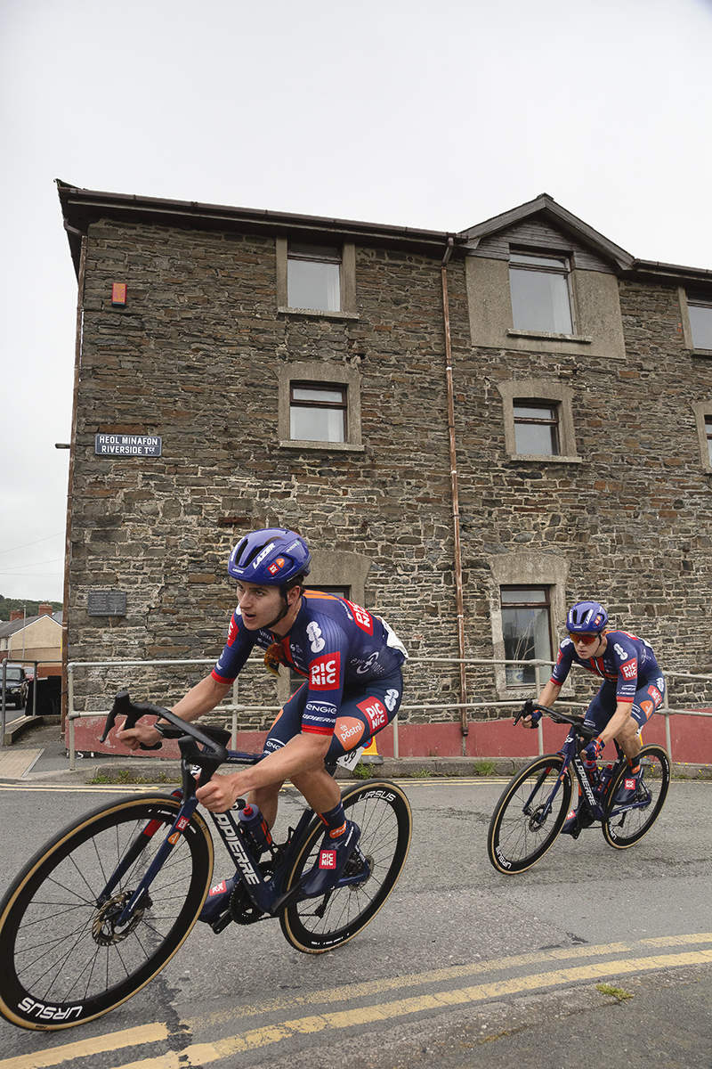 British National Road Championships 2025 - Road Race - Men - Jacob Bush & Angus Stoneham of Development Team Picnic PostNL round a corner past a stone building in  Aberystwyth