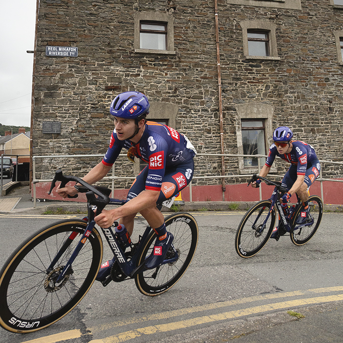 British National Road Championships 2025 - Road Race - Men - Jacob Bush & Angus Stoneham of Development Team Picnic PostNL round a corner past a stone building in  Aberystwyth