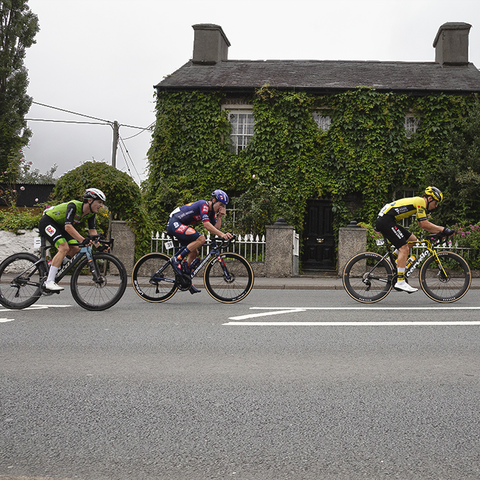British National Road Championships 2025 - Road Race - Men - a group of riders race past a stone cottage covered in ivy in Llanfarian