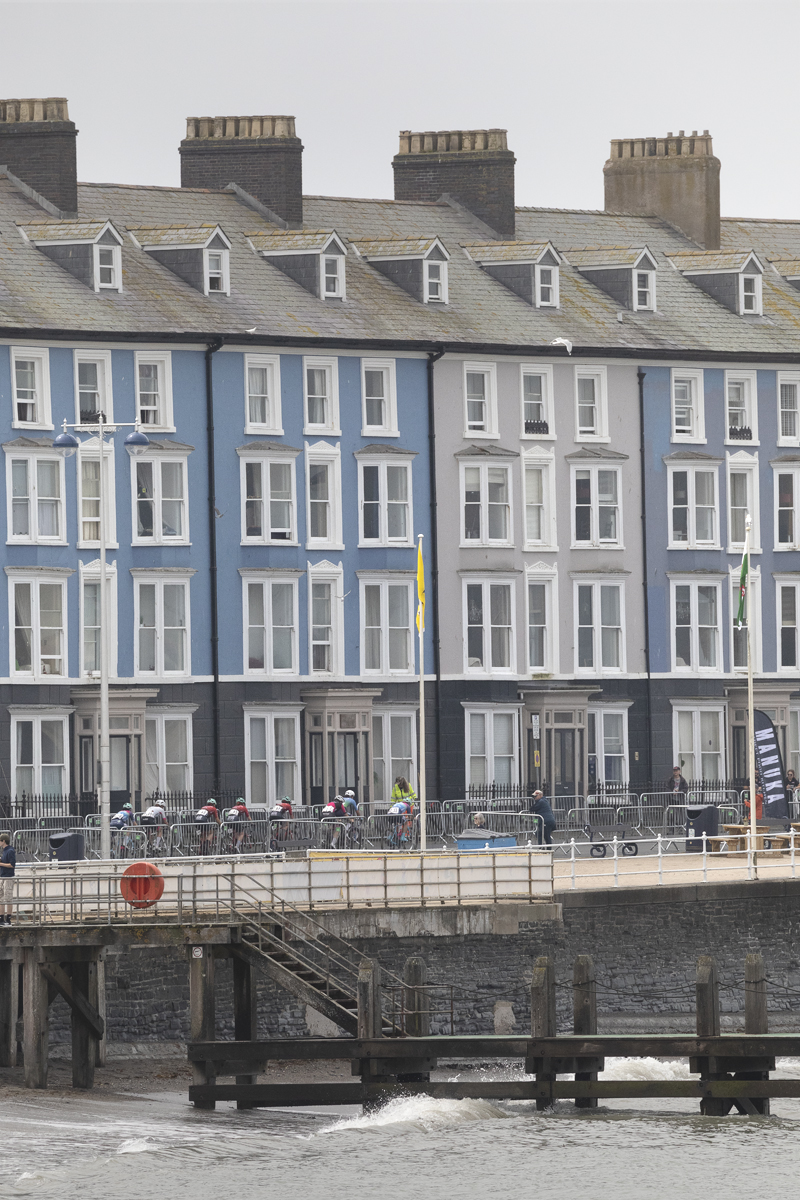 British National Road Championships 2025 - Road Race - Women - Aberystwyth - a group of riders rides past buildings painted in differing shades of blue on the seafront with waves crashing in the foreground