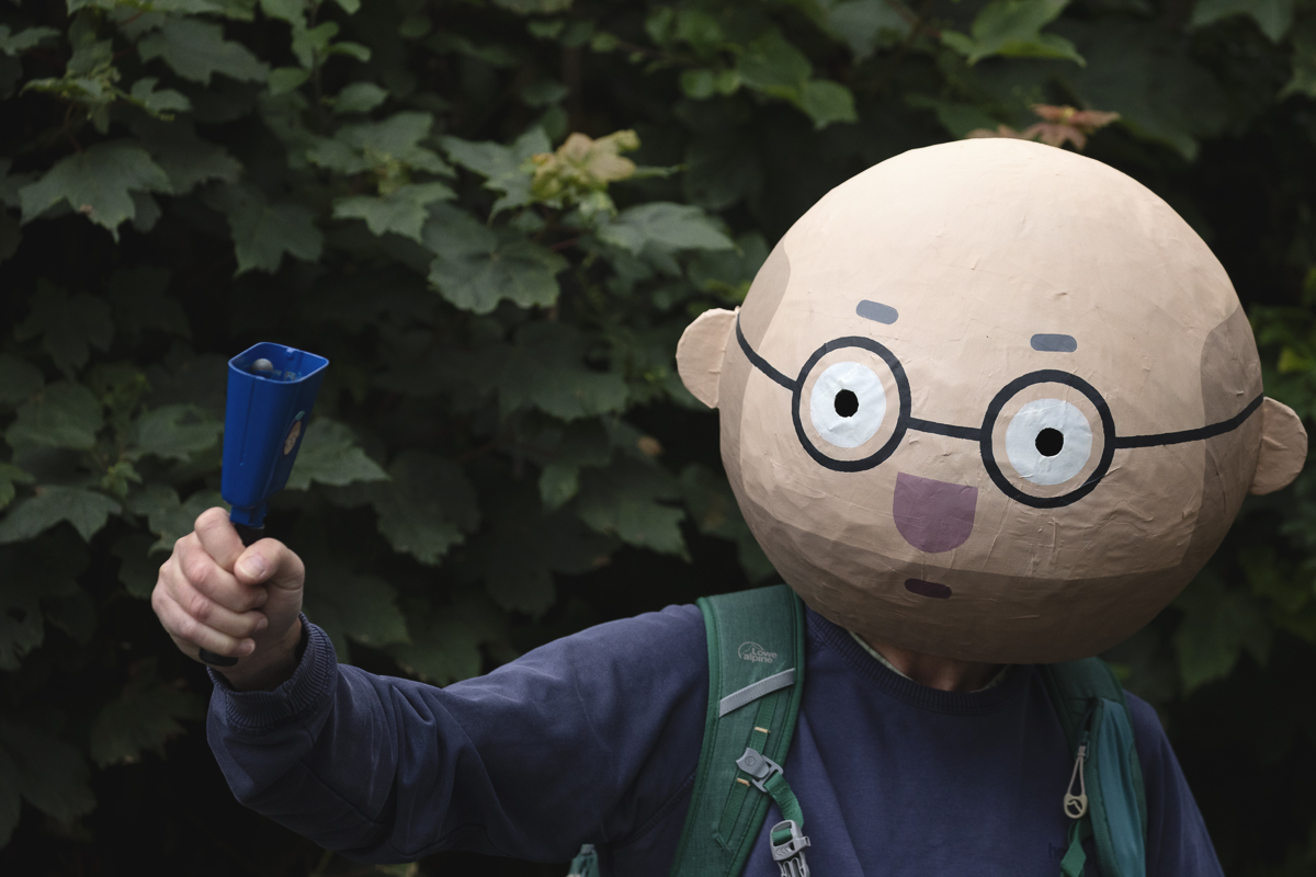 British National Road Championships 2025 - Time Trial - U23 Men - a fan wears a large Papier-mâché head and rings a cow bell to encourage riders