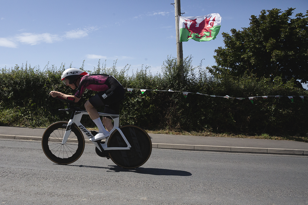 British National Road Championships 2025 - Time Trial - Elite Men - Ben Pease of Moonglu SpatzWear in a tuck position as he races past a Welsh flag blowing in the breeze