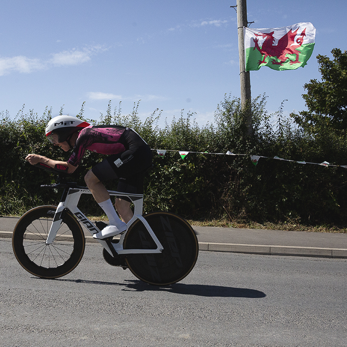 British National Road Championships 2025 - Time Trial - Elite Men - Ben Pease of Moonglu SpatzWear in a tuck position as he races past a Welsh flag blowing in the breeze