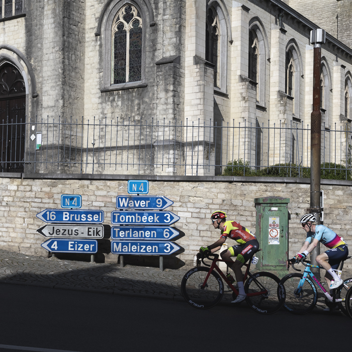 De Brabantse Pijl 2024 - Riders pass road signs to Jezus-Eik outside the church in Overijse