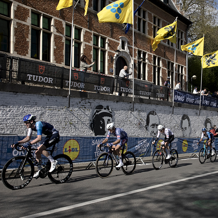 De Brabantse Pijl - Riders take on the S-Bocht Overijse past a white wall covered in the portraits of past cycling legends