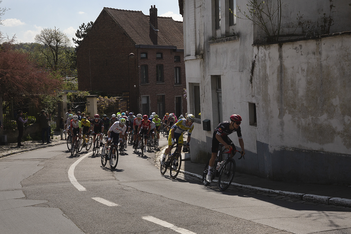 De Brabantse Pijl 2025  - A line of riders round the corner on the climb past a run down whitewashed building