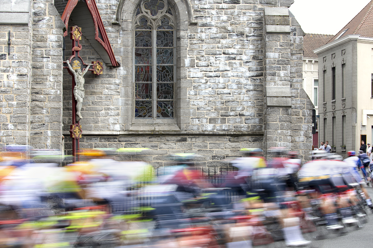 Dwars Door Vlaanderen 2022 - The peloton rushes past a church and depiction of Jesus on the cross