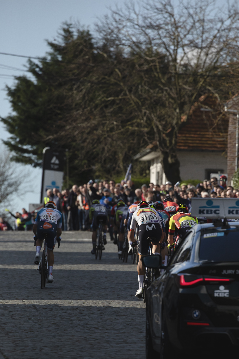 Dwars Door Vlaanderen 2024 - Riders seen from behind followed by a race vehicle up the Nokereberg