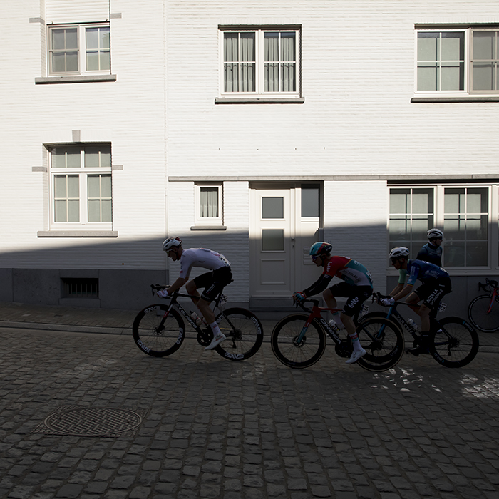 Dwars Door Vlaanderen 2024 - A group of riders in shadow against a bright white washed frontage of a building