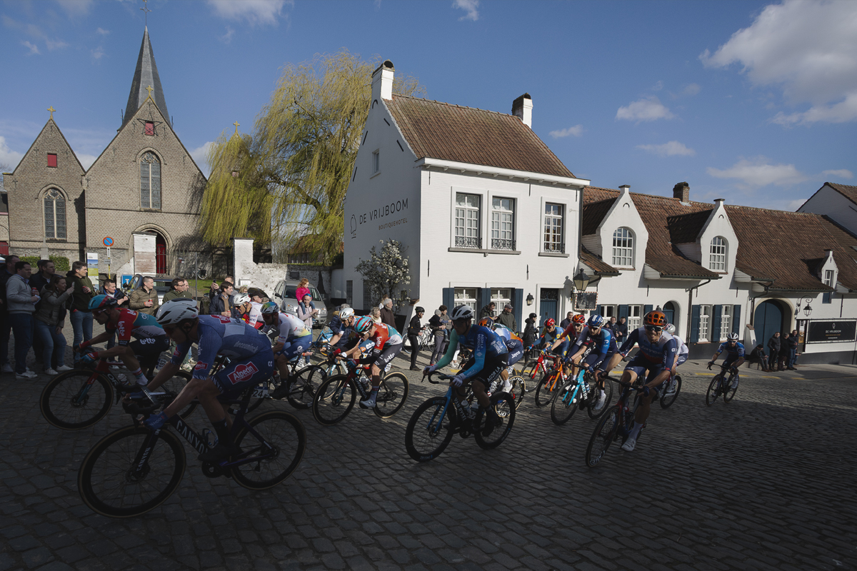 Dwars Door Vlaanderen 2024 - Riders pass through the main square in Nokere, white washed buildings and a church behind them