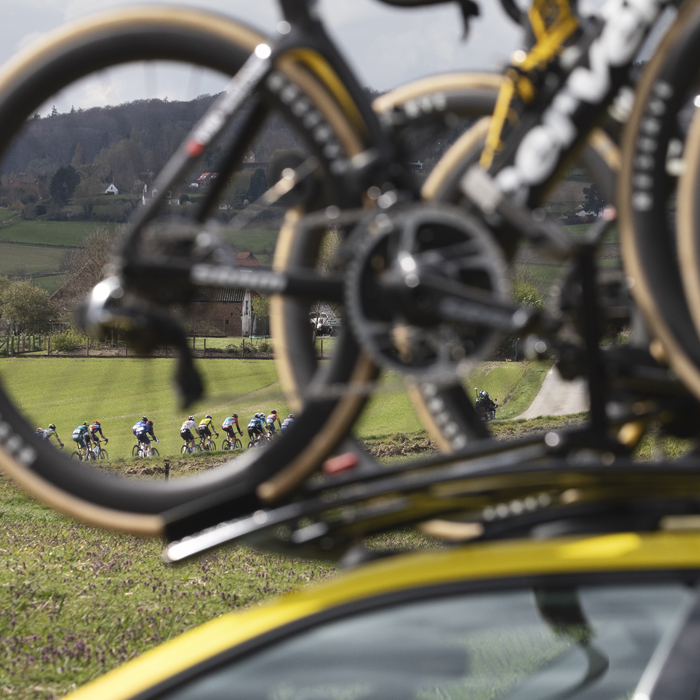 Dwars Door Vlaanderen 2024 - A group of riders on Rue des Chapelles are framed by the wheel of a bike on a team car
