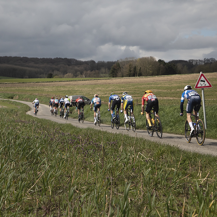 Dwars Door Vlaanderen 2024 - Riders pass by a slippery road sign on Rue des Chapelles