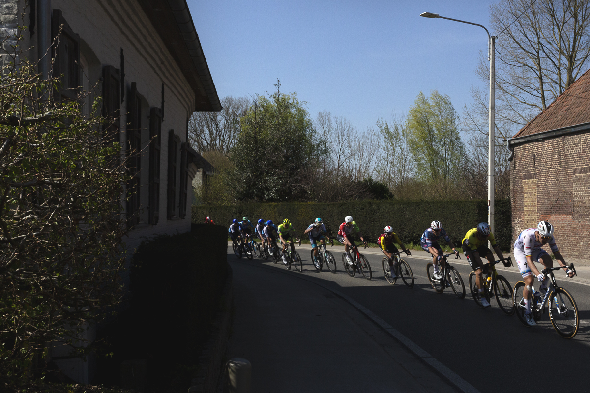 Dwars Door Vlaanderen 2025 - A strung out line of riders take a bend in the road past an old brick built barn