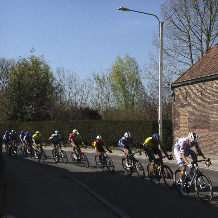 Dwars Door Vlaanderen 2025 - A strung out line of riders take a bend in the road past an old brick built barn