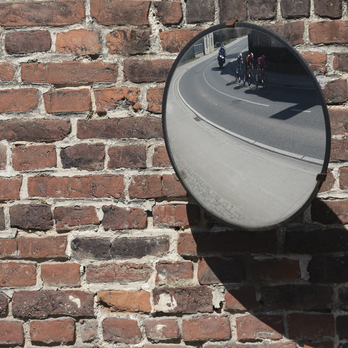 Dwars Door Vlaanderen 2025 - Riders reflected in a roadside mirror on a brick wall