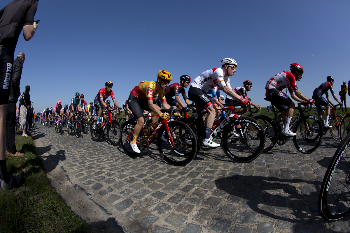 E3 Saxo Bank Classic 2022 - Long shadows are cast by fans at the side of the road as riders traverse the cobbles of Holleweg