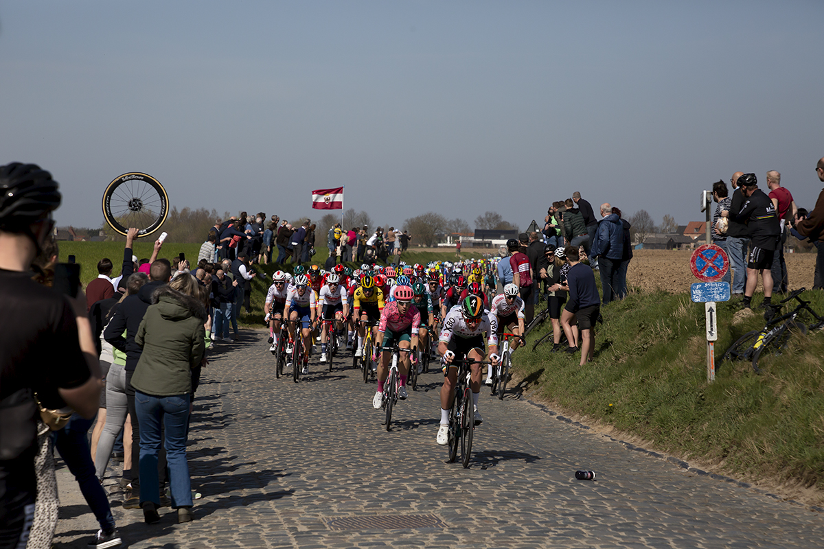 E3 Saxo Bank Classic 2022 - The peloton tackles the cobbles of Holleweg as a soigneur holds up a spare wheel at the side of the course