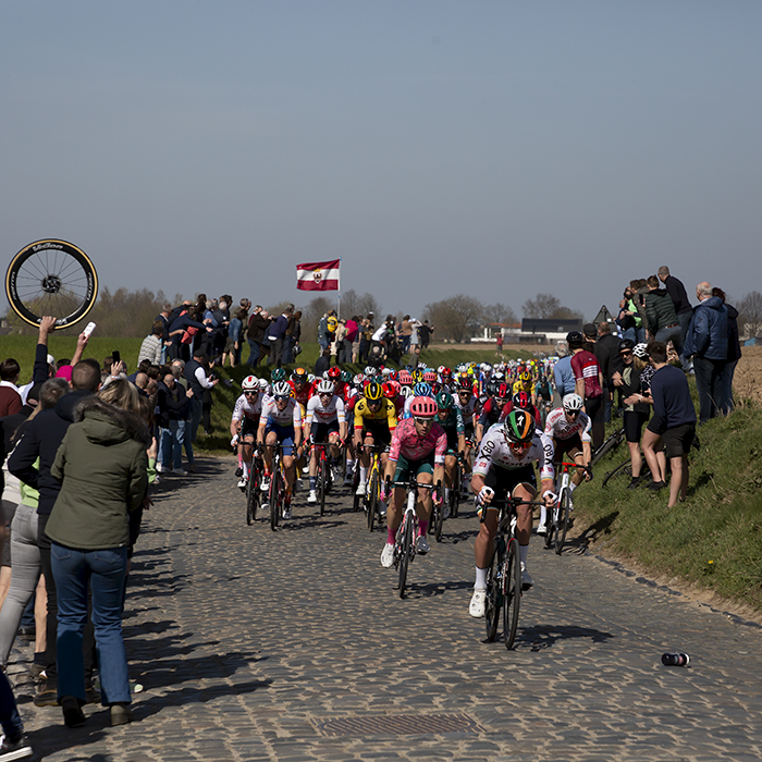 E3 Saxo Bank Classic 2022 - The peloton tackles the cobbles of Holleweg as a soigneur holds up a spare wheel at the side of the course