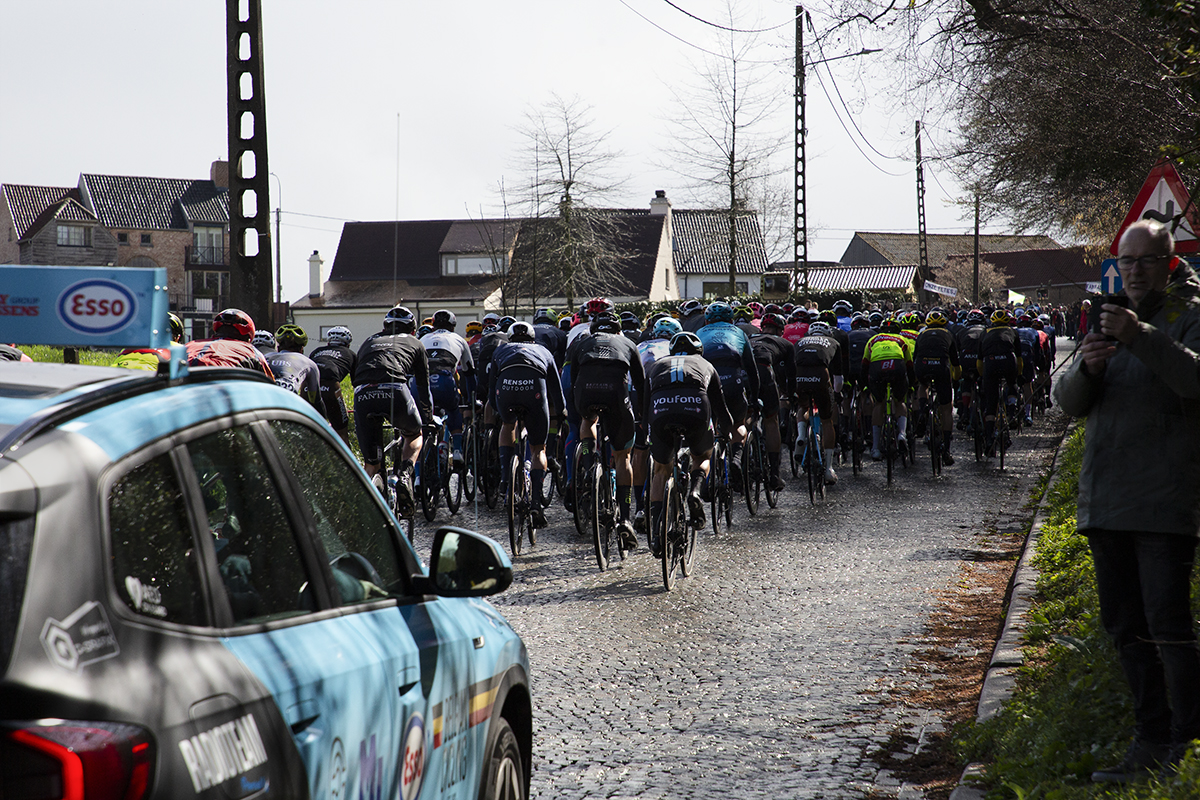 E3 Saxo Classic 2023 - The peloton shown riding away with a Belgian cycling branded car in the foreground