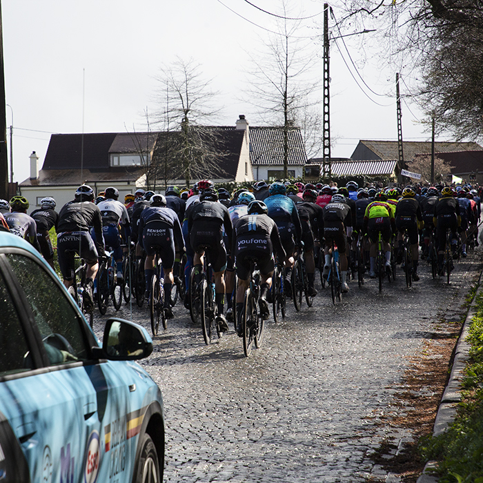 E3 Saxo Classic 2023 - The peloton shown riding away with a Belgian cycling branded car in the foreground