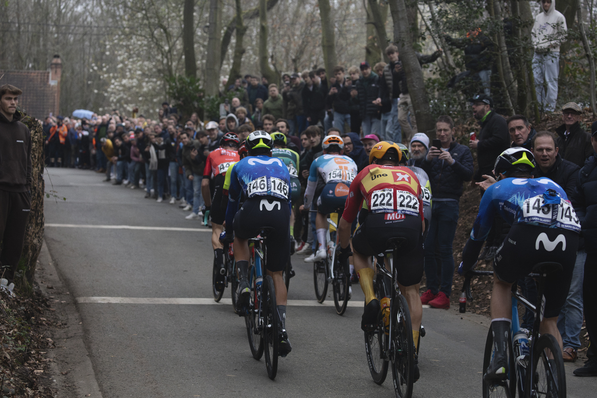 E3 Saxo Classic 2024 - Riders from behind on the climb as fans watch from raised vantage points at the side of the narrow road