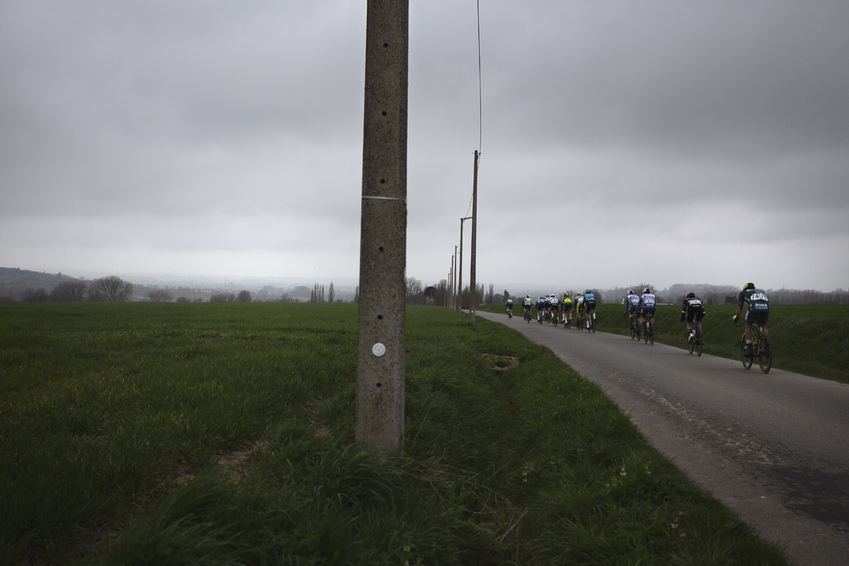 E3 Saxo Classic 2024 - Riders strung out down the road with conrete lamp posts to one side