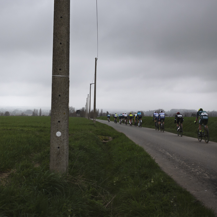 E3 Saxo Classic 2024 - Riders strung out down the road with conrete lamp posts to one side