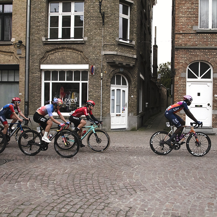 Gent Wevelgem Vrouwen 2024 - Riders from the side as they pass by the facades of tall brick buildings inIeper / Ypres